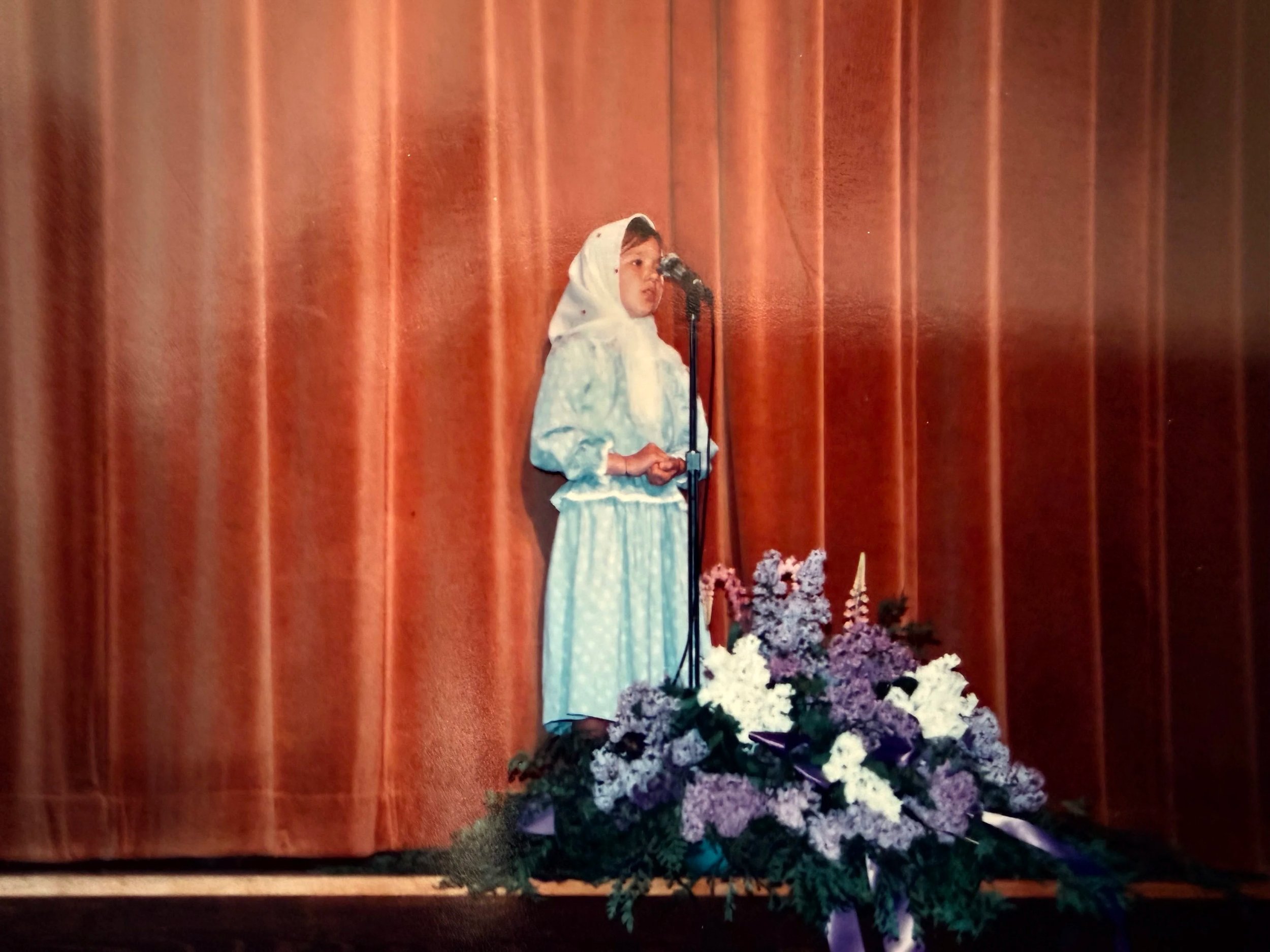 A young girl dressed in a light blue dress and white headscarf standing on stage with a microphone, in front of a closed red curtain, surrounded by flower arrangements.
