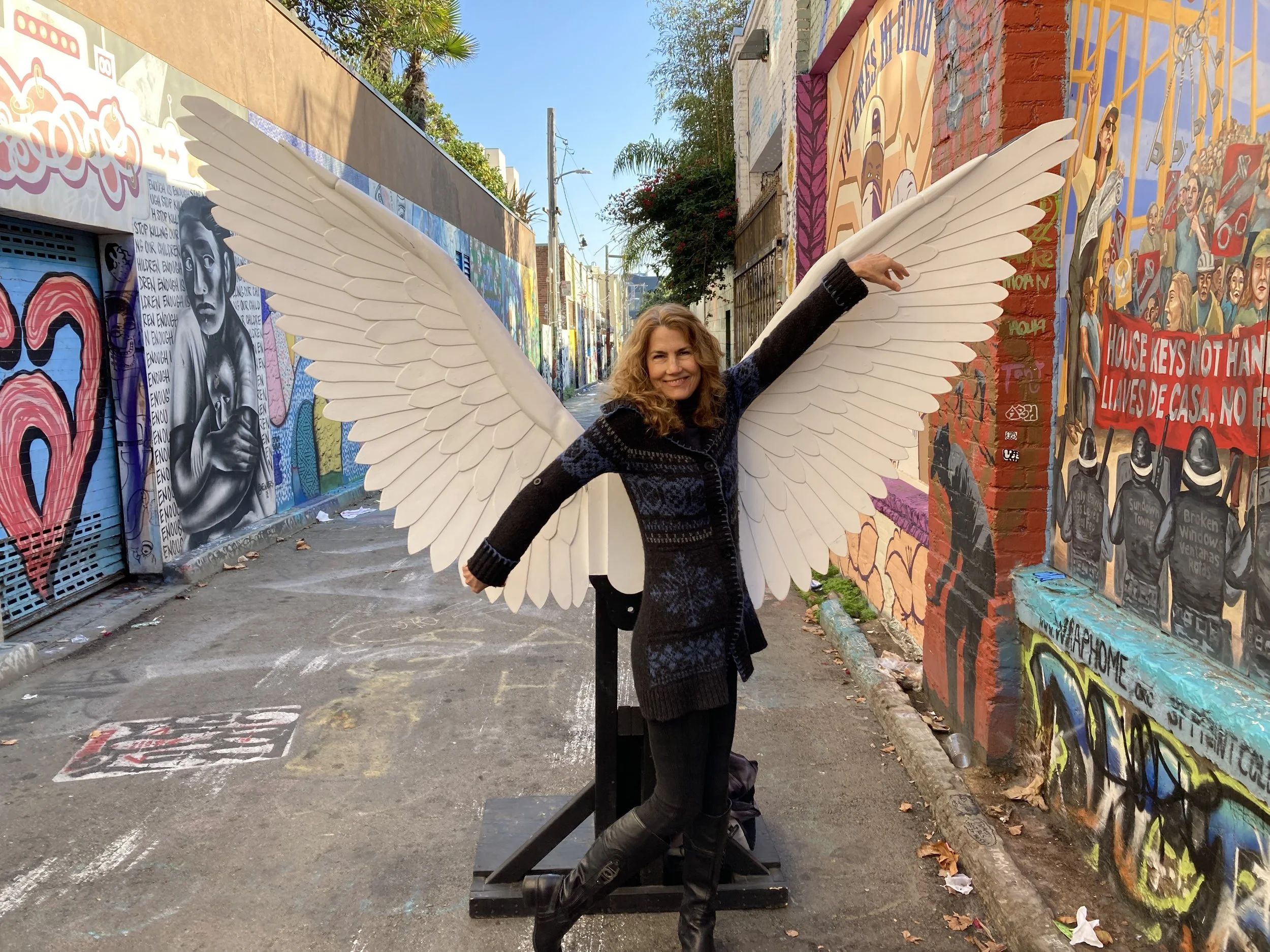 Woman with curly hair smiling and pointing to her angel wings costume, standing in front of colorful street murals.