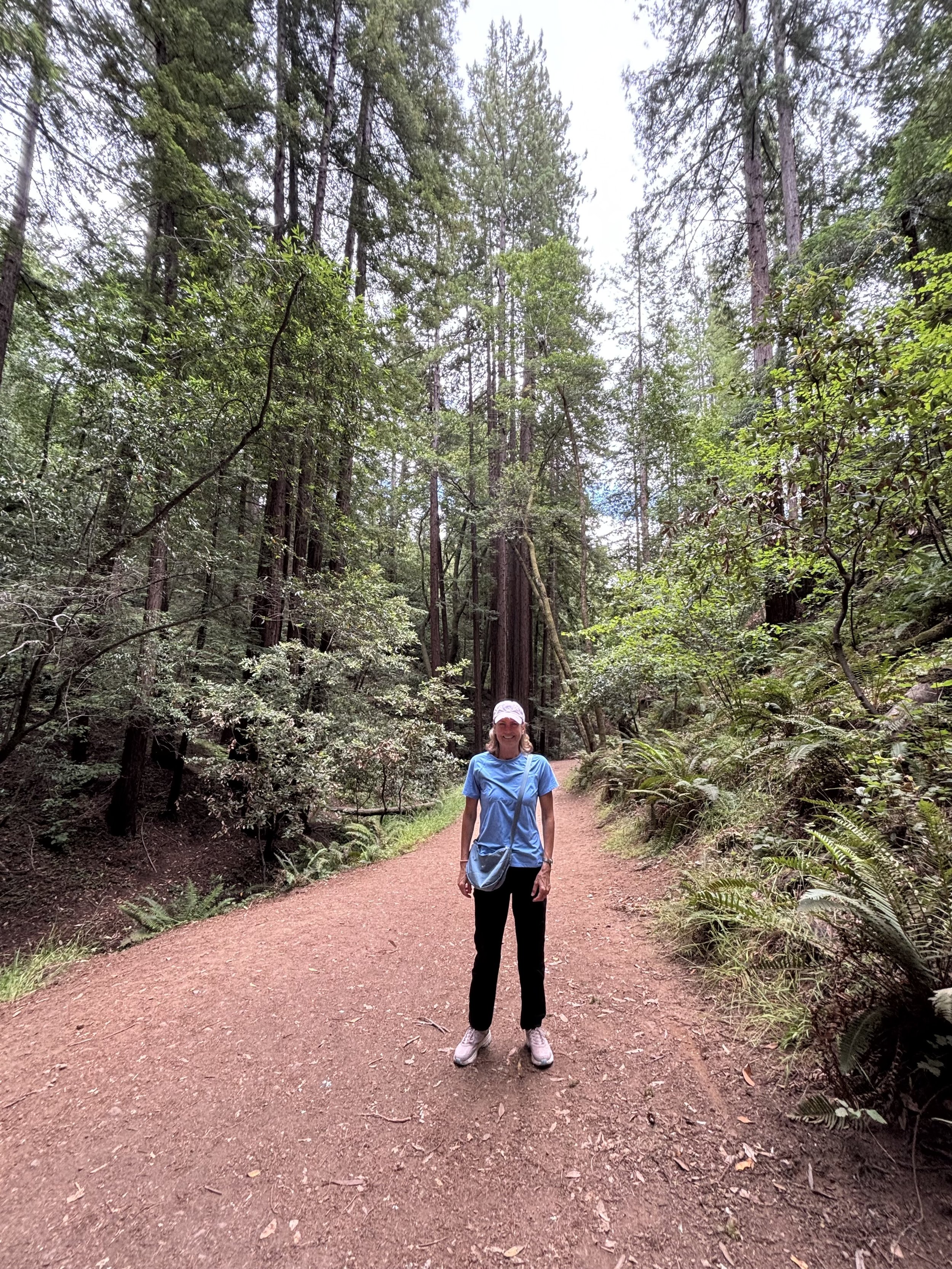 A woman in a blue shirt, black pants, and gray shoes standing on a dirt trail in a lush green forest, smiling at the camera.
