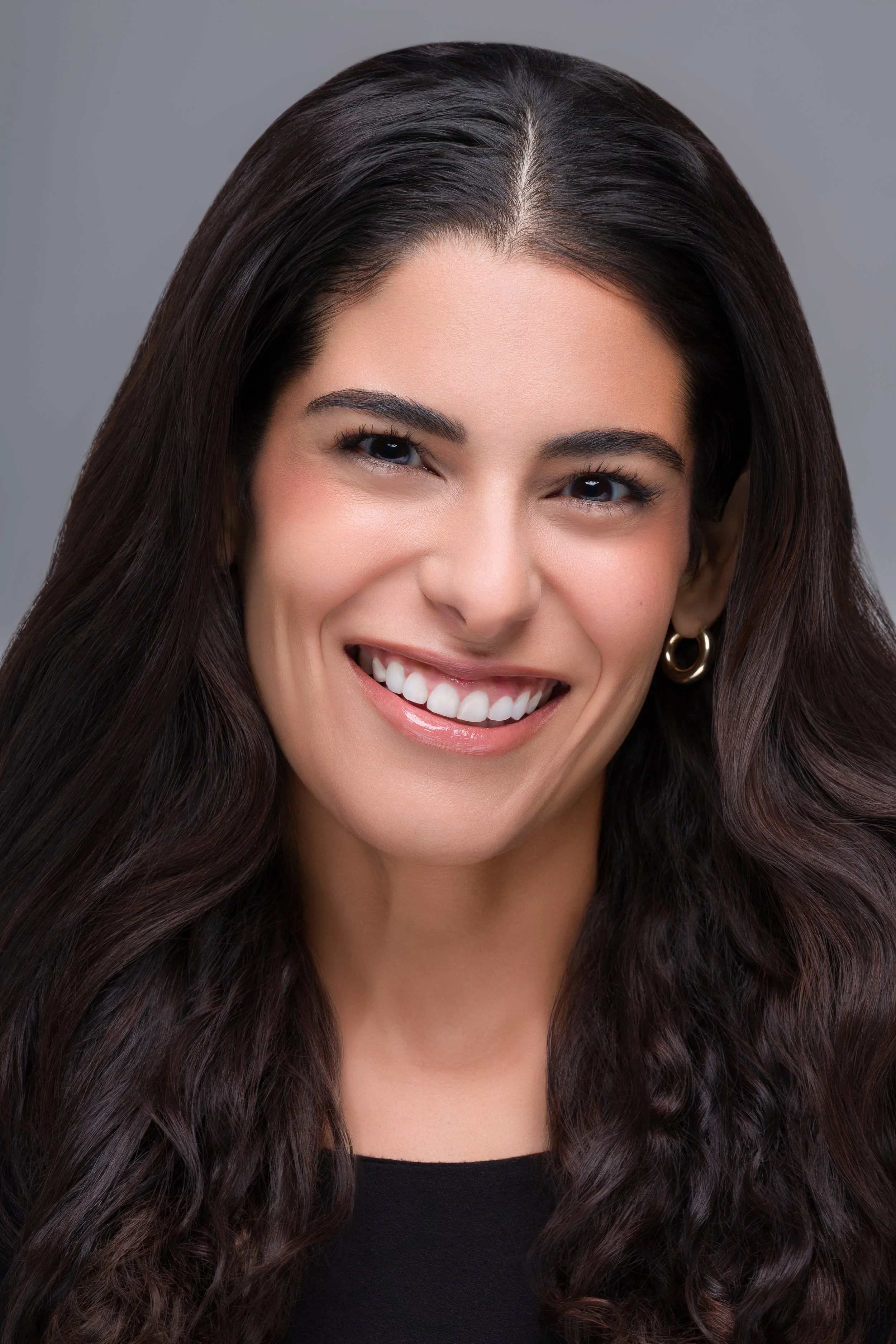 Close-up of a woman with long dark hair, smiling and showing her teeth, wearing a black top and gold hoop earrings.