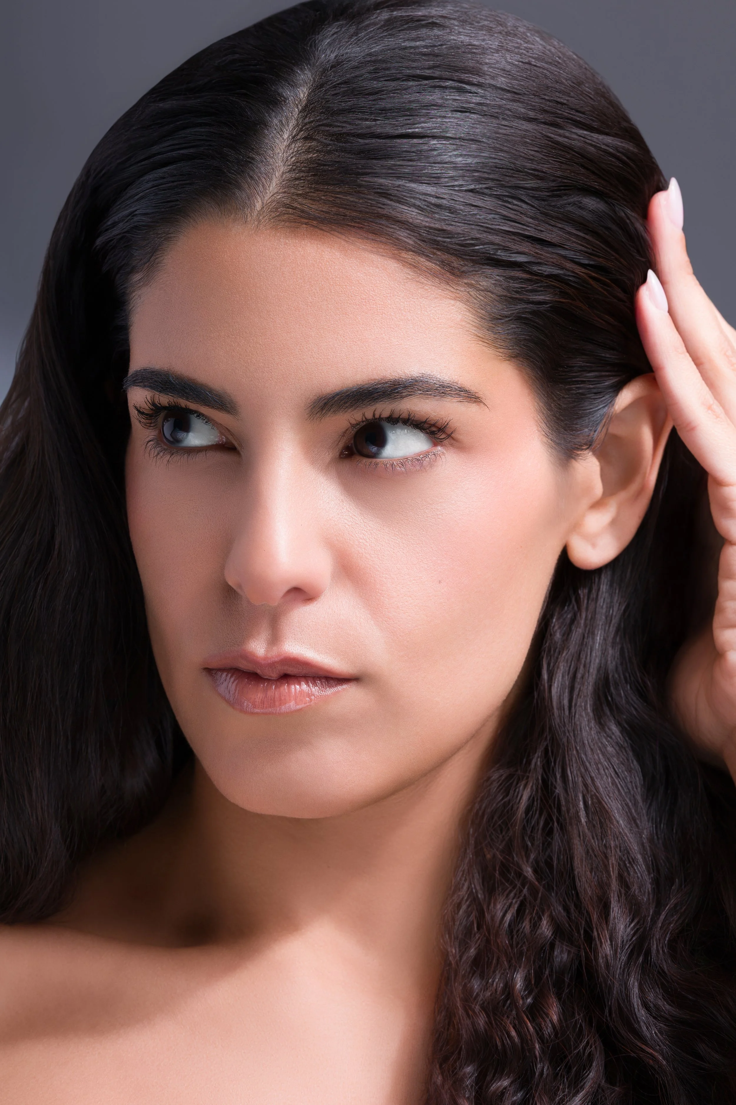 Close-up of a woman with long, dark wavy hair, lightly touching her hair with one hand, and looking off to the side.
