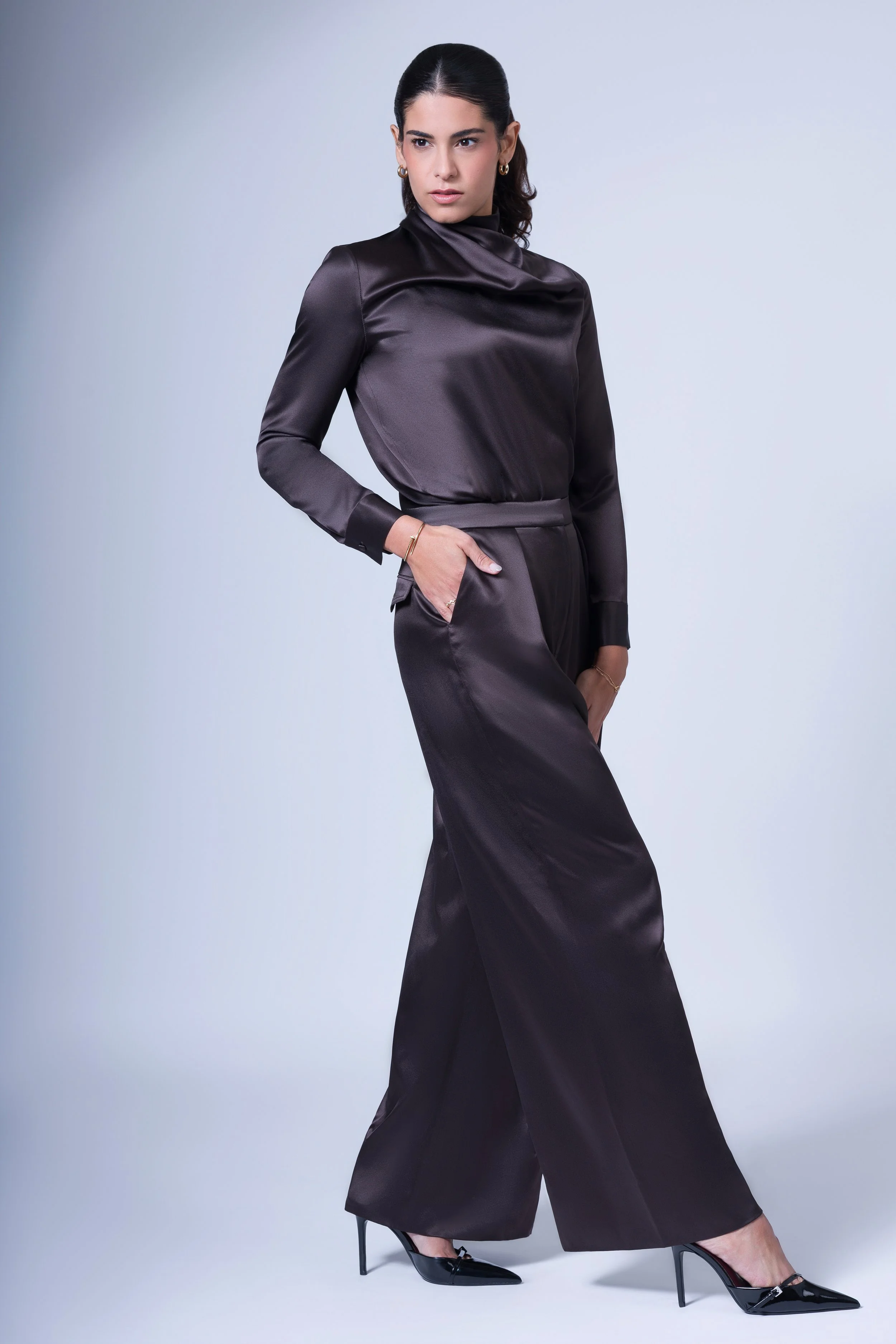 A woman with dark hair styled back, wearing a shiny black satin top and matching wide-leg satin pants, black high heels, and minimal jewelry, standing against a plain light gray background.