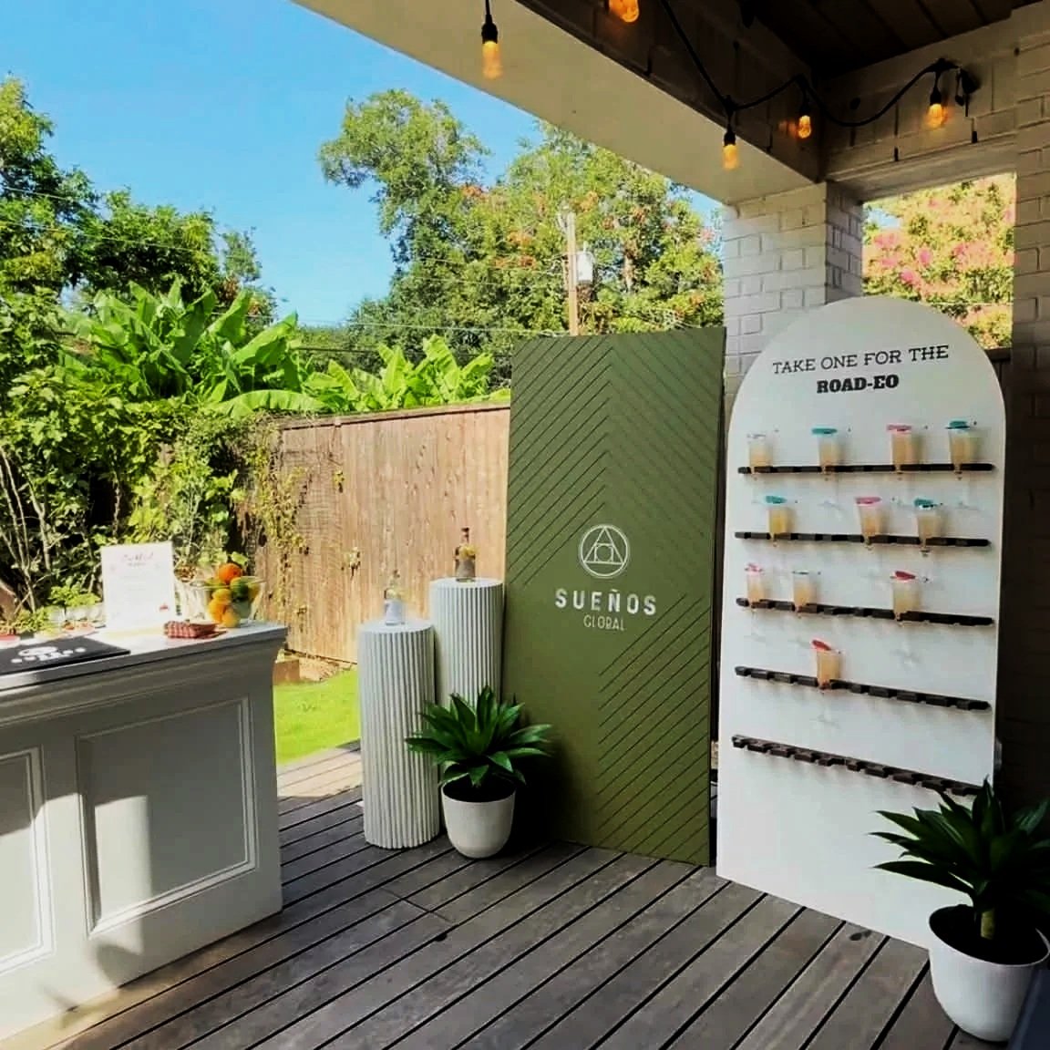 Outdoor patio scene with a white display stand holding cups of bubble tea, a green sign with the logo 'Sueños Global,' potted plants, and a wooden deck with string lights overhead.