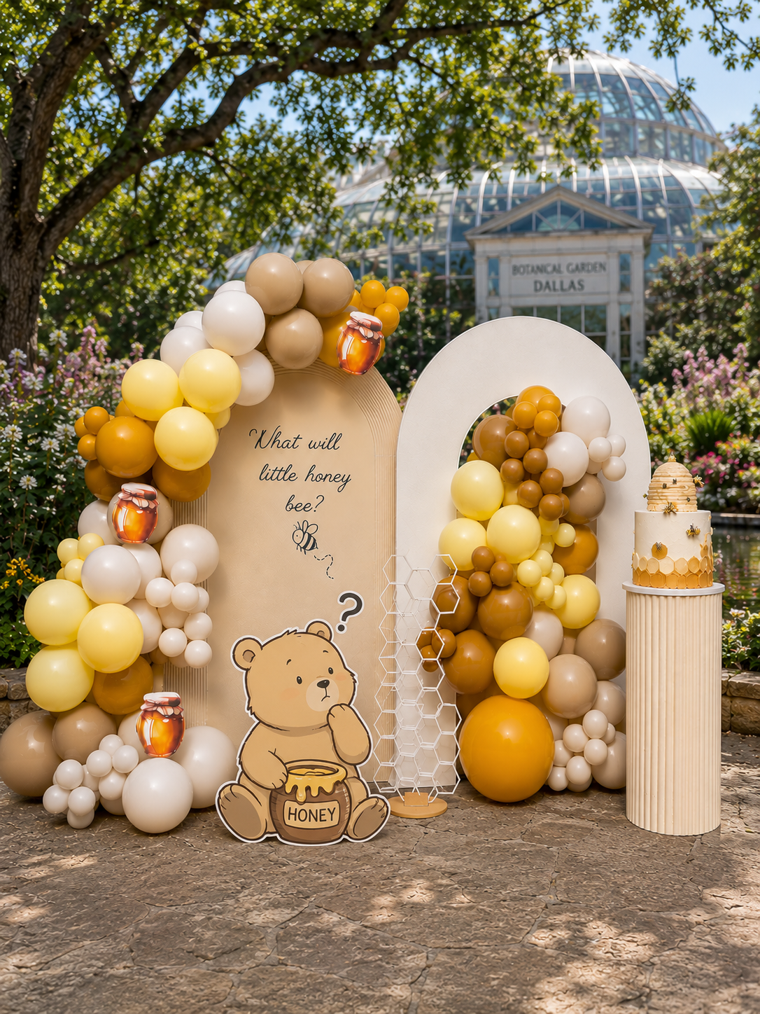 Decorative display at a botanical garden with balloons in shades of white, cream, yellow, and brown, a cartoon bear holding a honey jar with a question about honey bees, and a white cake with honey-themed decorations, set outdoors near trees and a greenhouse labeled 'Botanical Garden Dallas.'