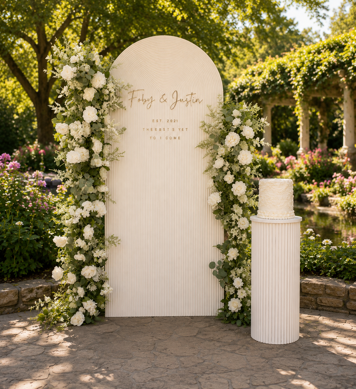 Wedding setup with a white floral arch, a white cake on a pedestal, and a sign for Foby and Justin surrounded by trees and flowers in outdoor garden setting.