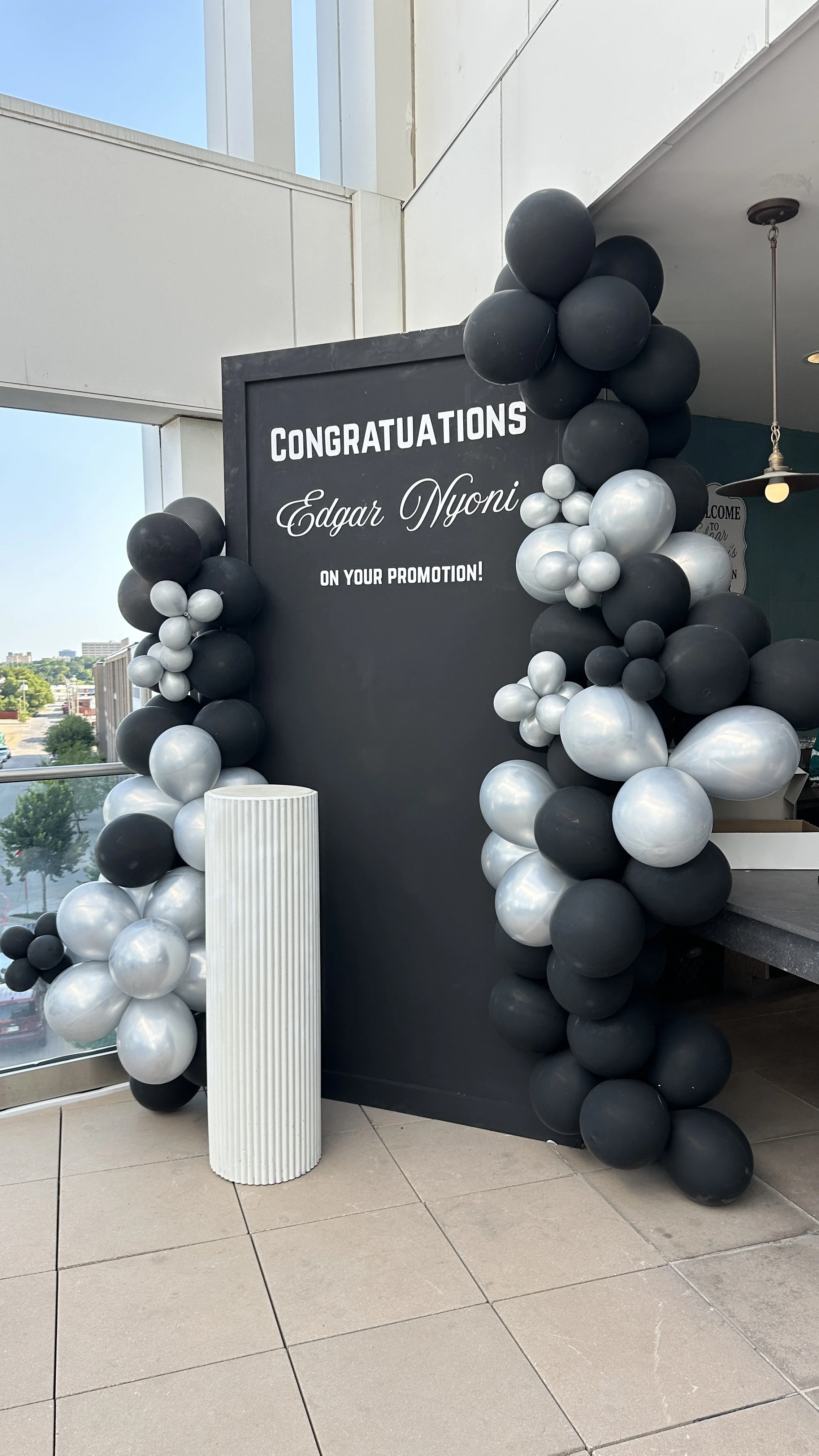 Black and silver balloon arch with a chalkboard sign celebrating Edgar Nyoni's promotion, on a balcony with city view.