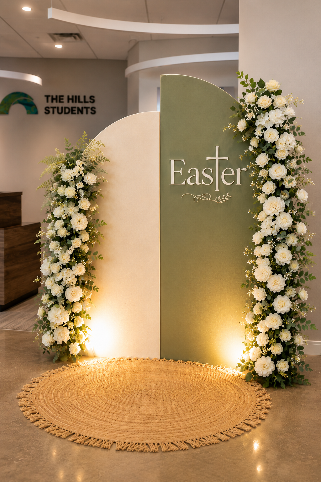 Easter themed floral display with white roses and greenery around a two-panel backdrop, illuminated from below, with a beige woven rug in the foreground.