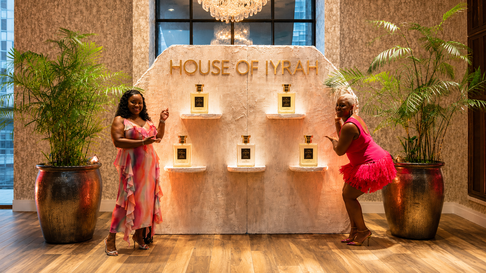 Luxury perfume product display featuring illuminated fragrance bottles, custom textured backdrop, gold lettering signage, tropical greenery, and event attendees at a high-end brand activation event.
