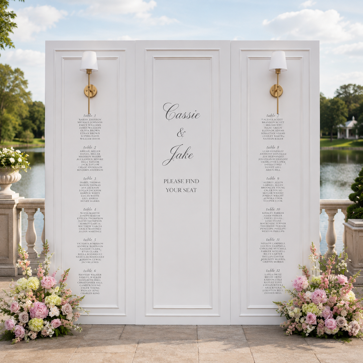 Wedding seating chart display with the names Cassie and Jake in the center, surrounded by floral arrangements of pink and white roses at the bottom left and right, and two wall sconces on either side against a lakeside outdoor setting with a balustrade and trees.