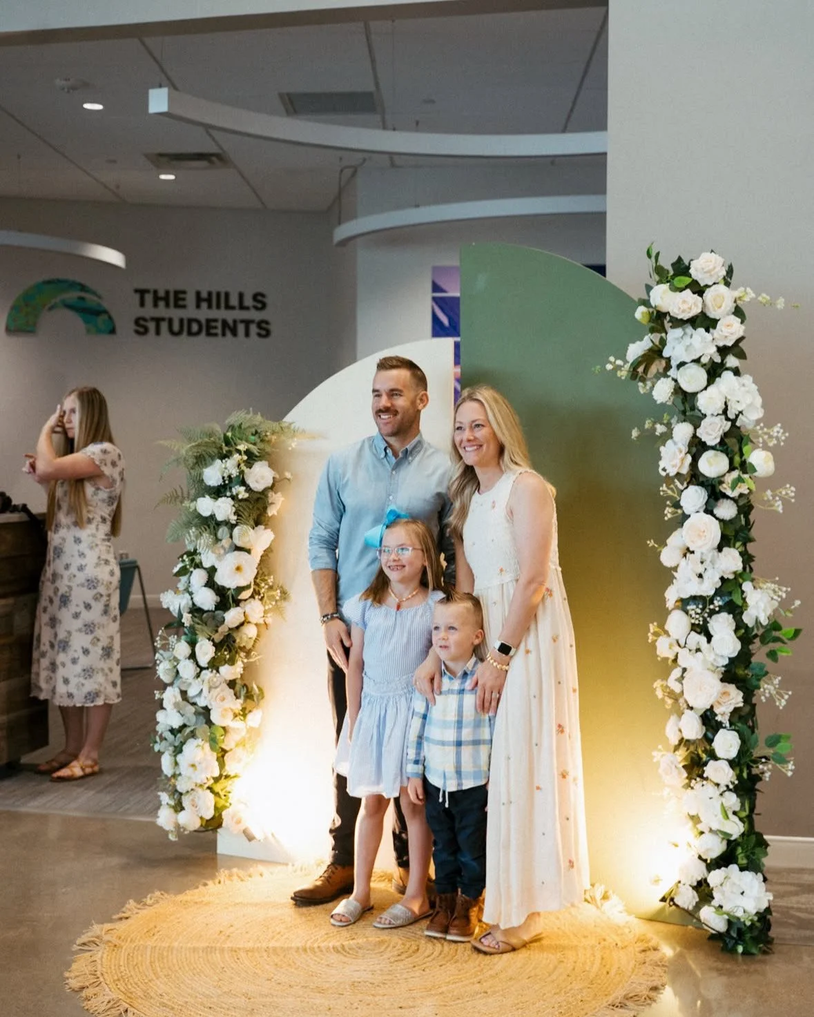 Family of four standing together and smiling at an indoor event, decorated with floral arrangements, with a backdrop lighting.
