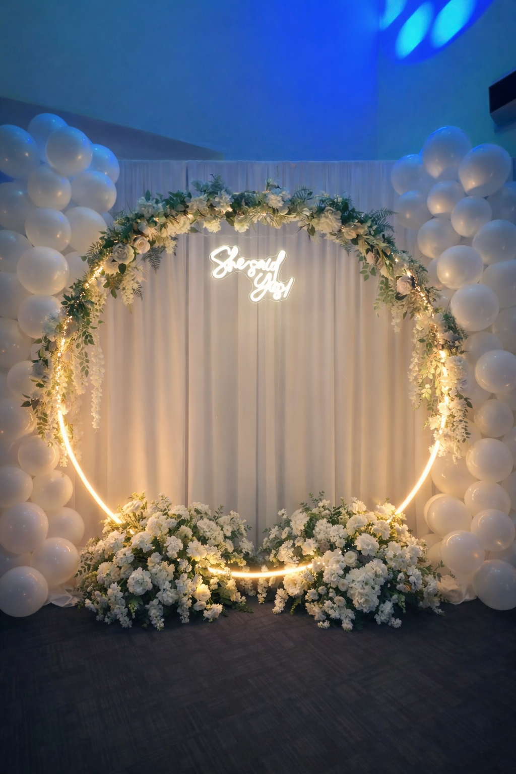 Elegant wedding arch decorated with white flowers, surrounded by white balloons and illuminated with warm lighting, with a backdrop of white curtains and a neon sign that reads 'She said yes'.