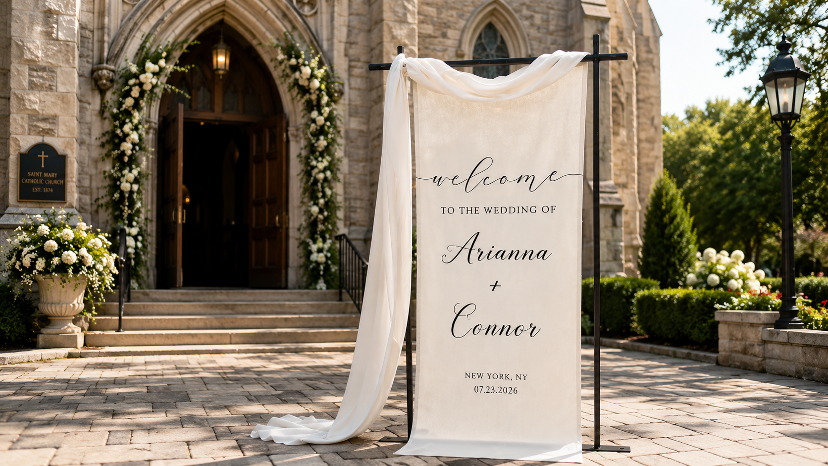 Elegant wedding welcome sign with flowing ivory fabric displayed in front of a stone church entrance decorated with white floral arrangements and greenery.