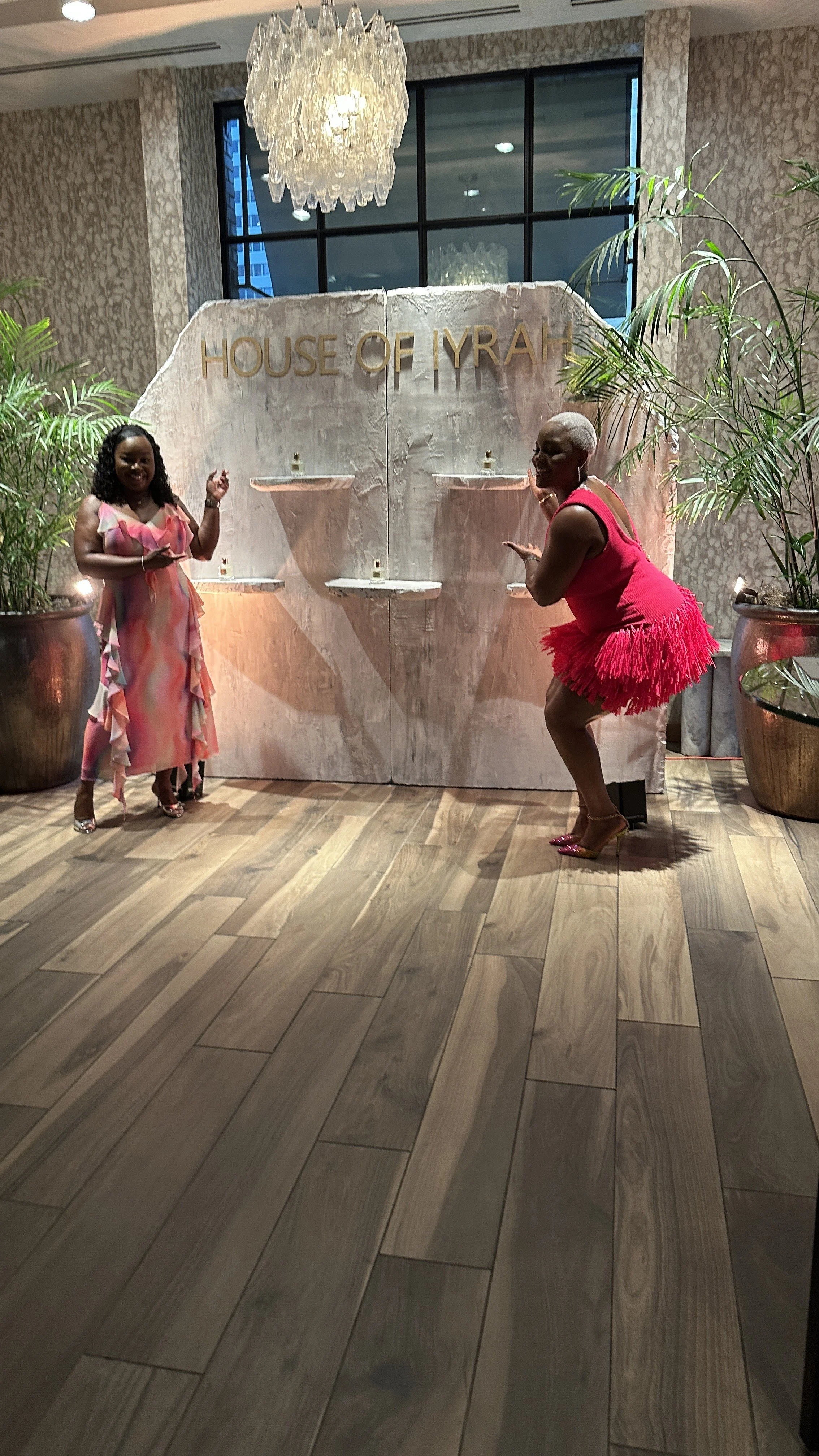 Two women standing in front of a backdrop that says 'House of IYAH' at an indoor event. One woman is smiling and wearing a colorful dress; the other woman is in a bright pink dress with a feathery skirt, posing playfully. Large potted plants are on either side of the backdrop.