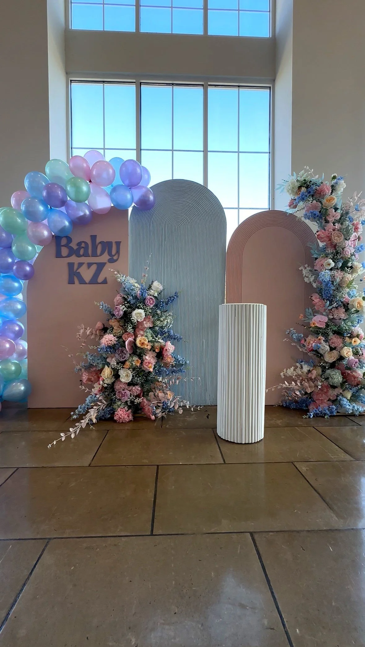 Decorative backdrop with pastel balloons, floral arrangements, and sign that says "Baby KZ" for a baby shower event.