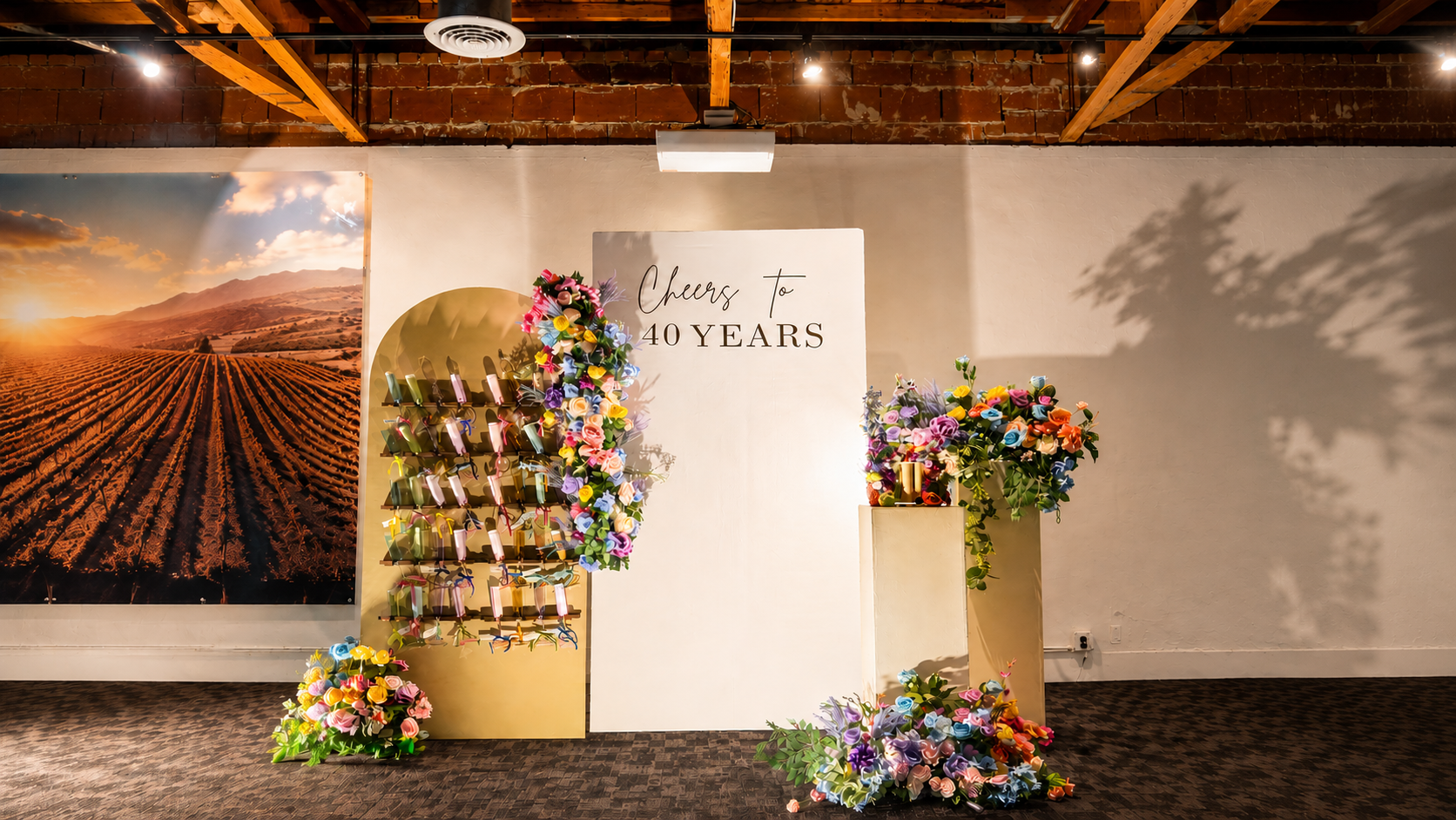 Decorative display celebrating a 40th anniversary with a large floral arrangement, a sign reading "Cheers to 40 Years," and a gold-colored stand with hanging gifts or favors. The background features a large mural of a vineyard at sunset and an exposed brick ceiling.