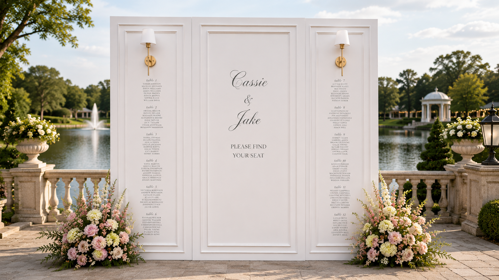 Elegant white wedding seating chart wall featuring custom paneling, floral arrangements, and gold sconces overlooking a lakeside wedding venue with stone terrace and garden views.
