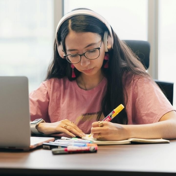 Woman wearing headphones writing notes beside laptop.