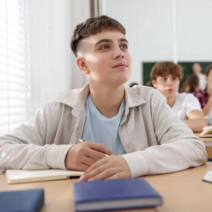Student holding a pen and looking thoughtful while seated at a desk in a classroom.