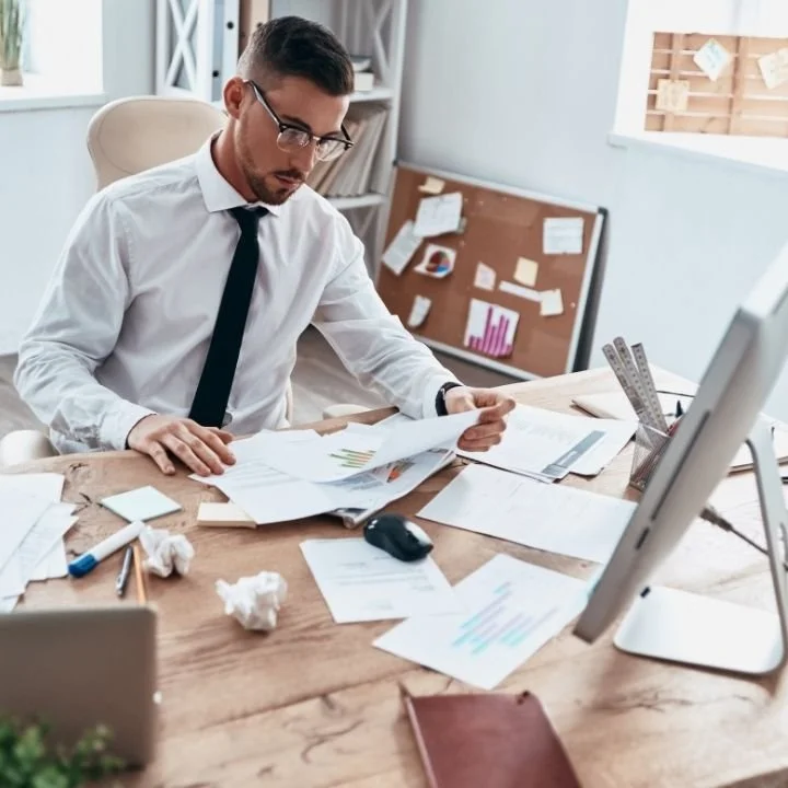 Adult man reviewing paperwork at desk, focused but visibly overloaded.