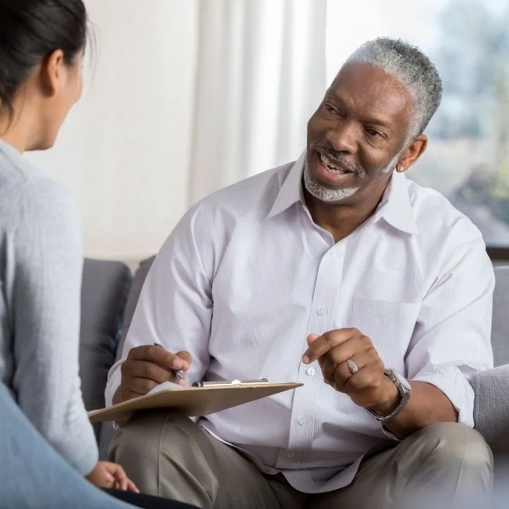 Therapist leans forward with a clipboard, speaking calmly while making eye contact as a client listens attentively.