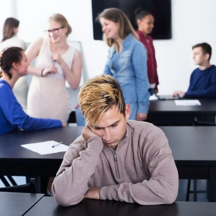 Teenager sitting alone in a classroom, looking sad and frustrated while other students talk in the background