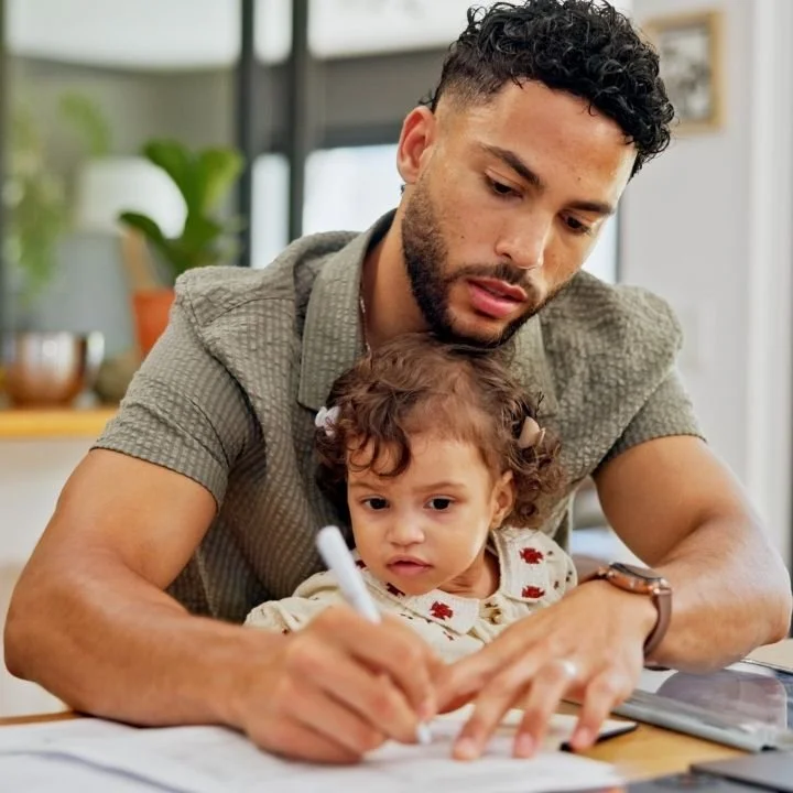 Father completing paperwork at home with his young daughter sitting on his lap.