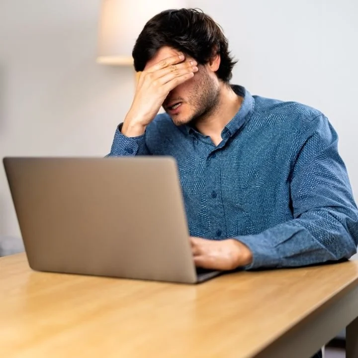 Adult man rubbing forehead while working on laptop at desk.