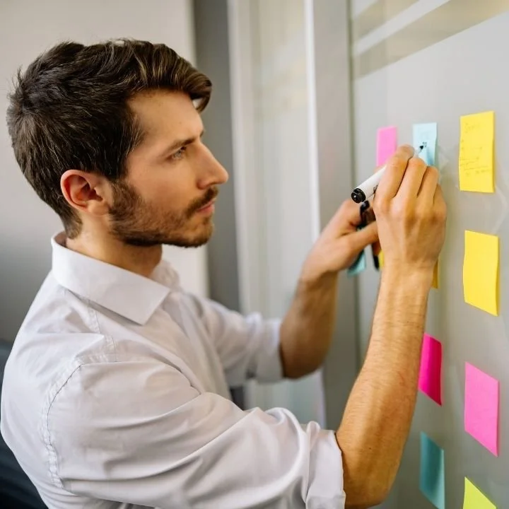 Man organizing colorful sticky notes on a board while managing tasks and priorities.