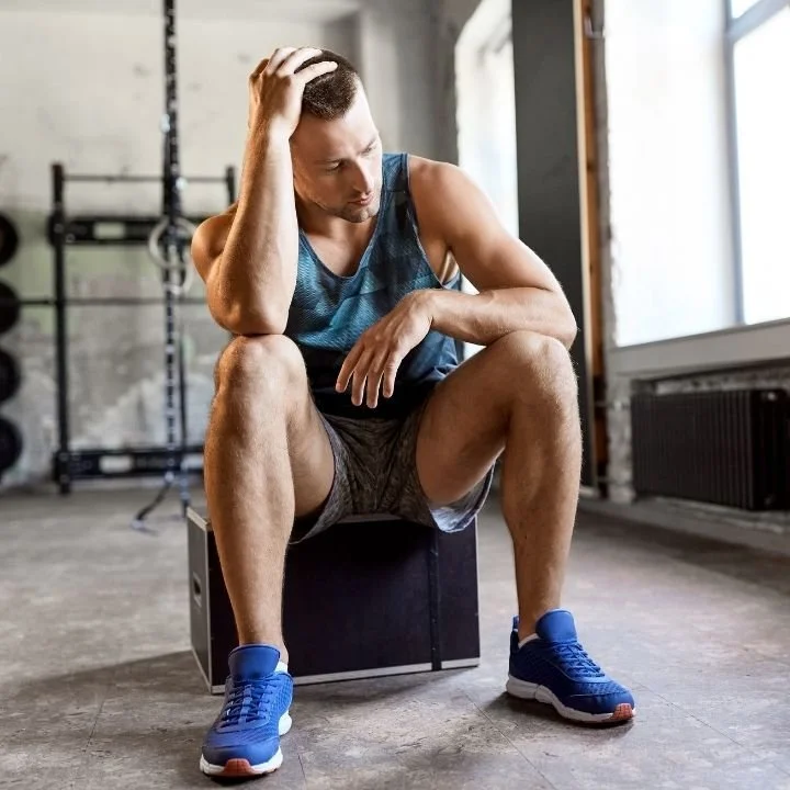 Man in workout clothes sitting on a box in a gym, looking down with his hand on his head