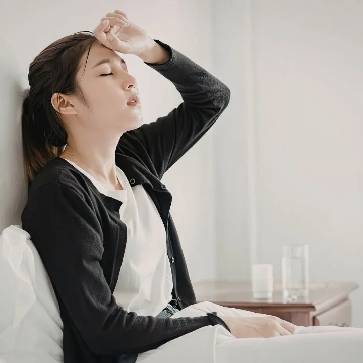 Woman sitting on a bed with her hand on her forehead while experiencing stress and fatigue.