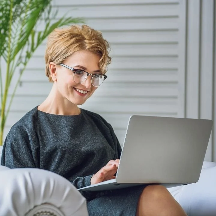 Woman smiling while typing on a laptop and sitting comfortably on a couch at home.