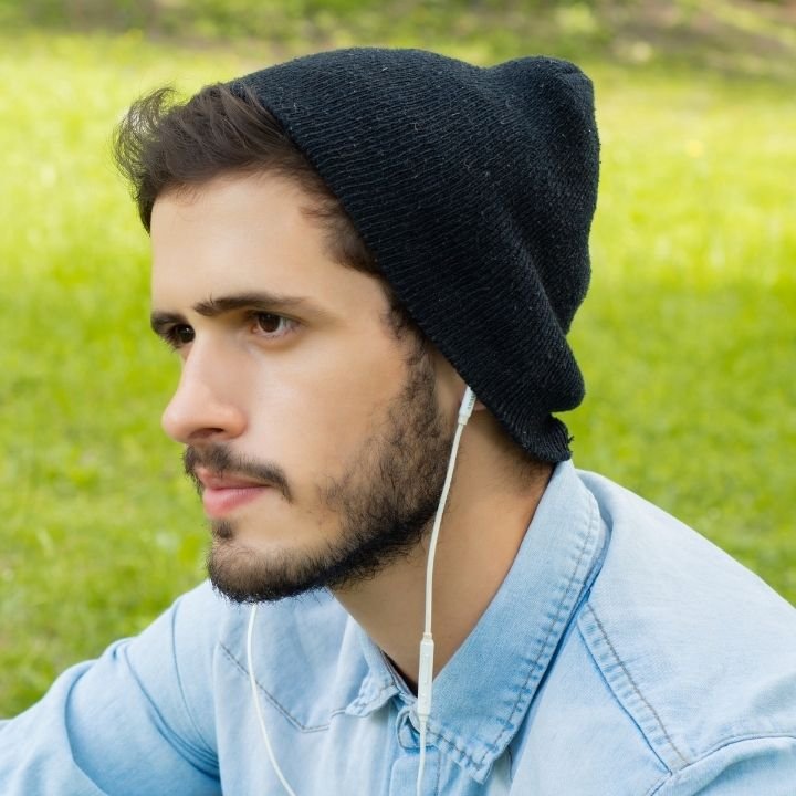 Man wearing beanie and earphones sitting outdoors looking thoughtful.