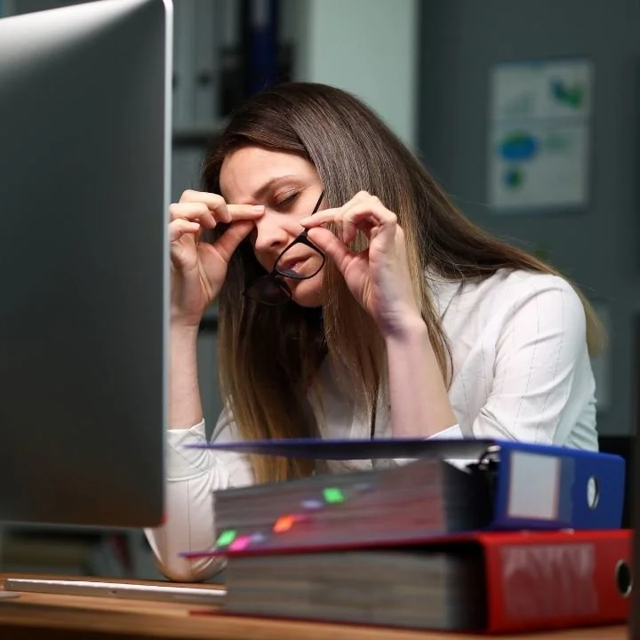 Woman at office desk rubbing her eyes while working on computer, showing signs of fatigue.