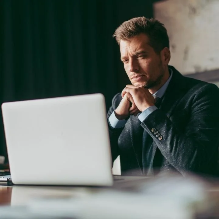 Man in dark suit looking intently at laptop screen in dim office