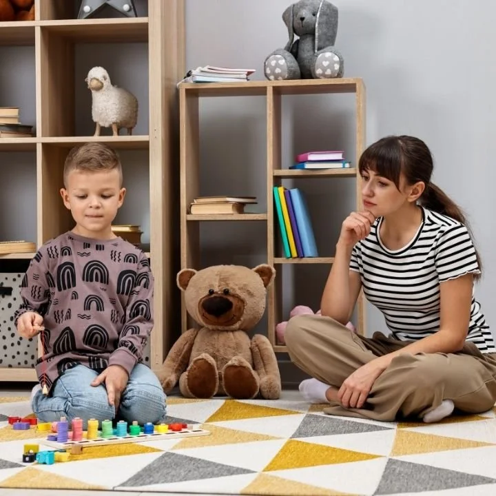 Child playing with blocks while therapist observes thoughtfully nearby.