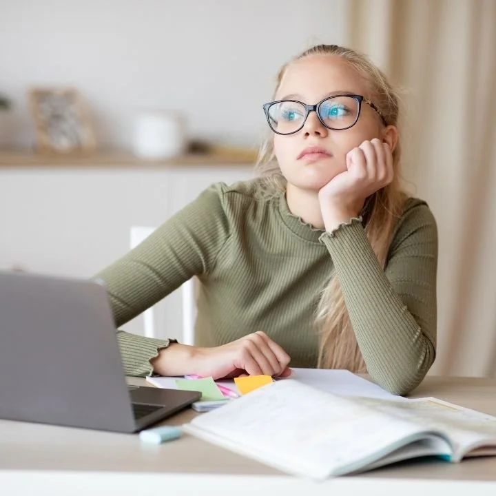 Female teen studying at desk, appearing distracted while working on laptop..