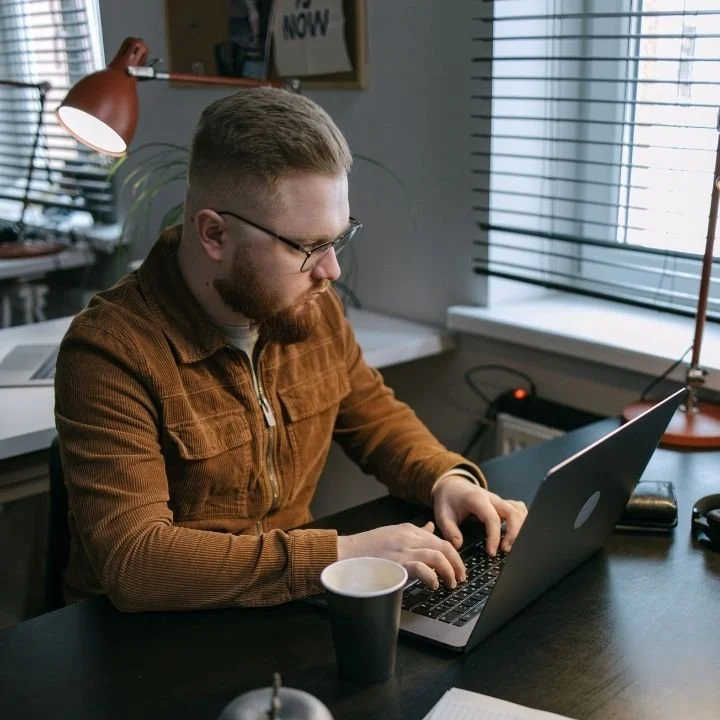 Man types on a laptop at a desk, leaning forward with focused attention.
