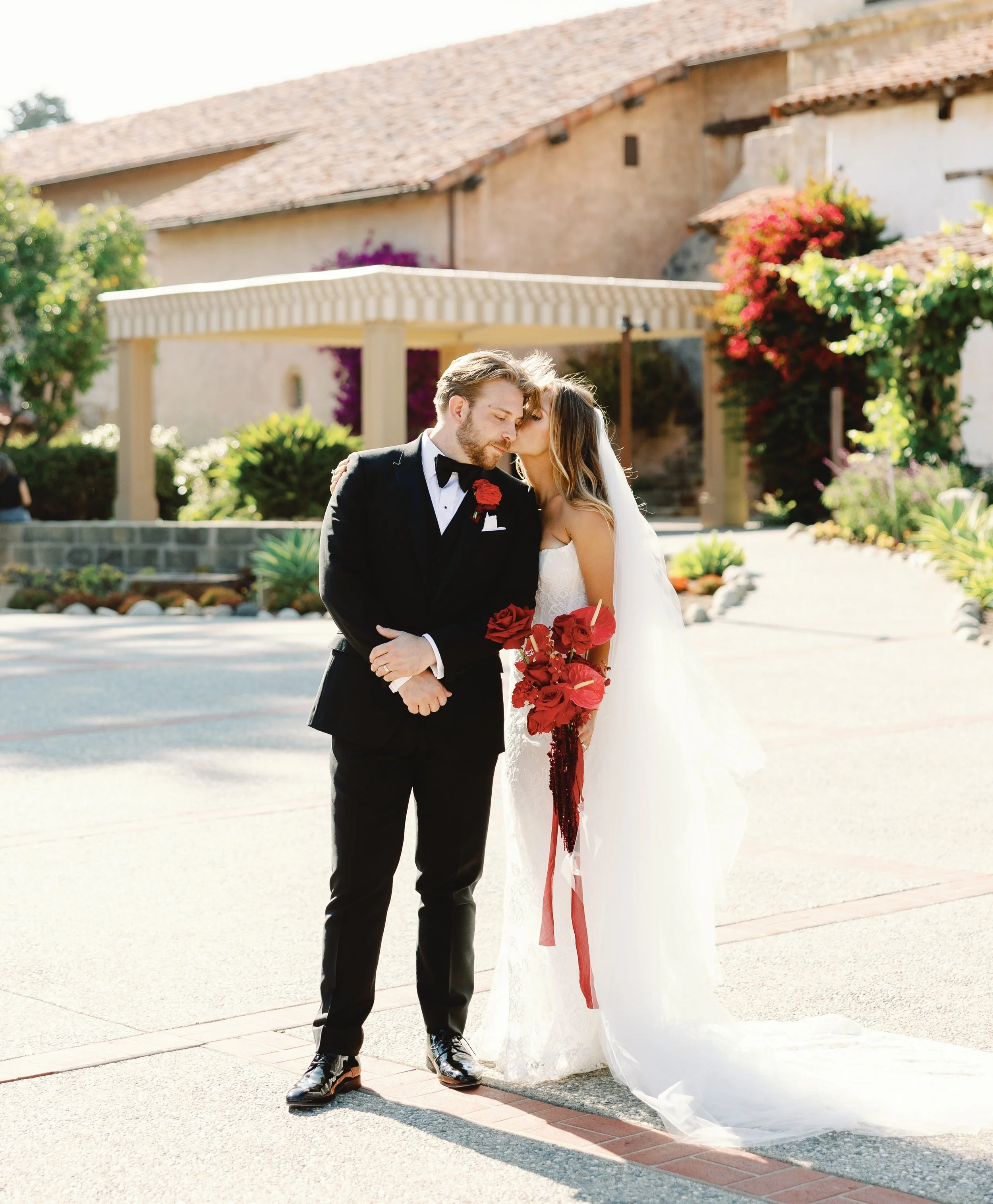 Bride and groom share an intimate kiss on the cheek outside a Spanish-style venue, bride holding a cascading red rose bouquet.