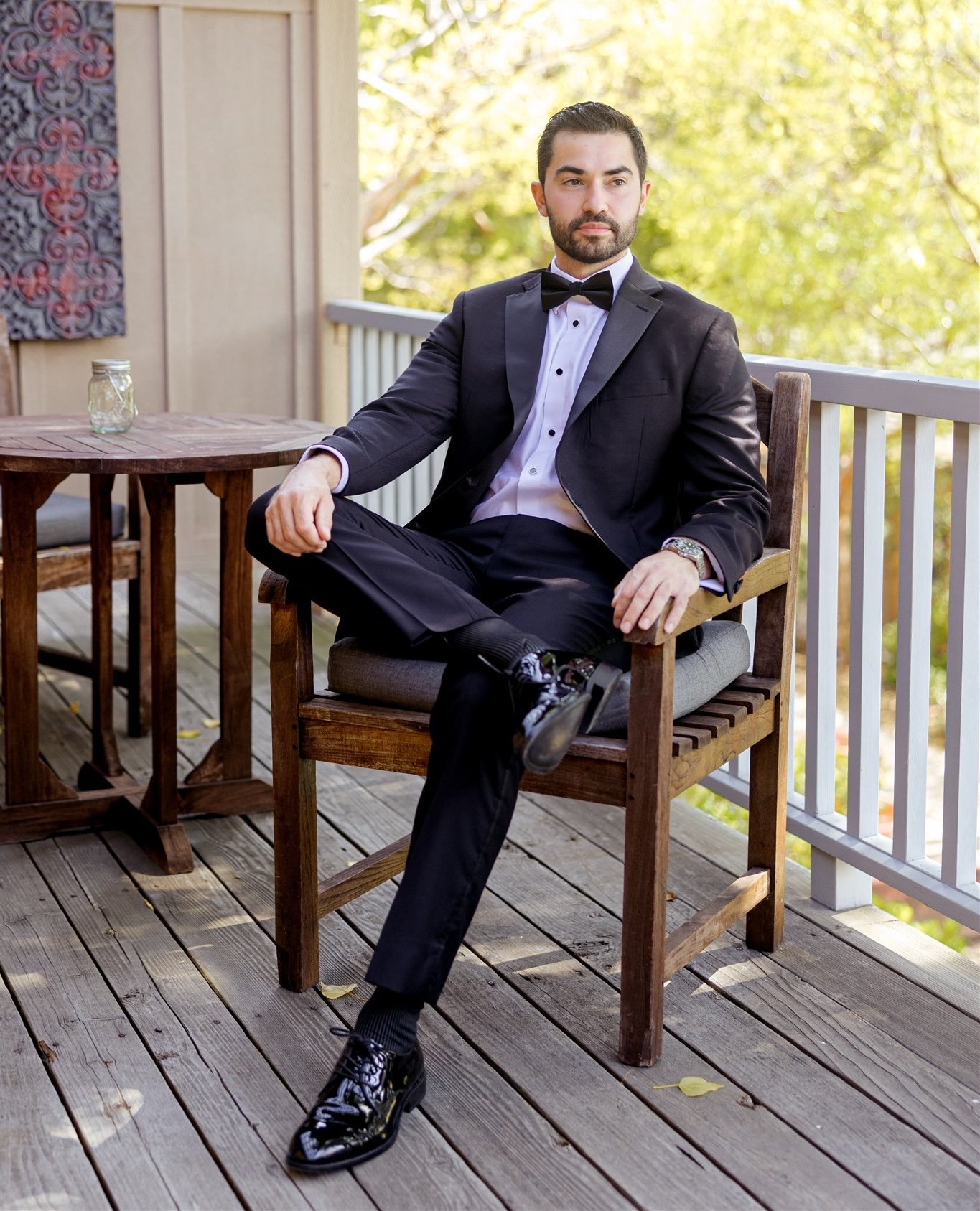 A well-dressed groom with a short dark beard and neatly styled hair sits relaxed on a wooden outdoor deck.
