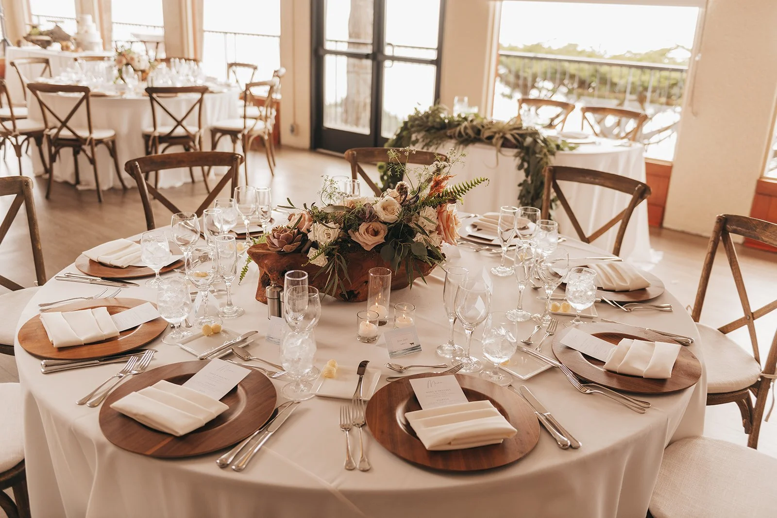 Wedding reception table with a rustic floral centerpiece, wooden chargers, and cross-back chairs in a sunlit room.