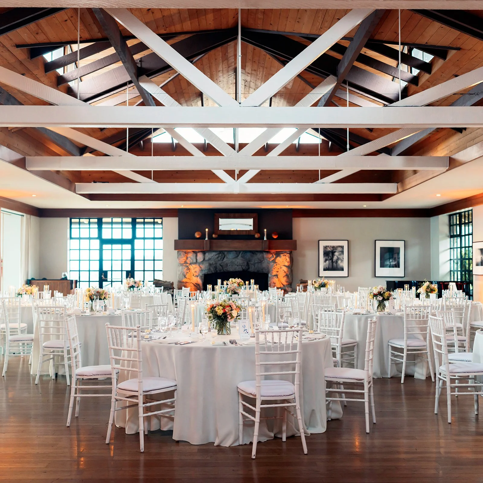 Elegant wedding reception at Highlands Inn featuring round tables, white Chiavari chairs, floral centerpieces, and a grand stone fireplace.
