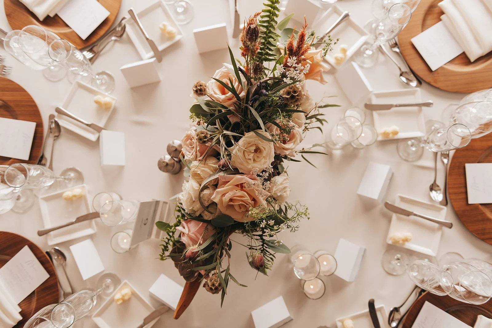 Overhead view of a wedding table setting with wooden charger plates and a blush rose and fern centerpiece.