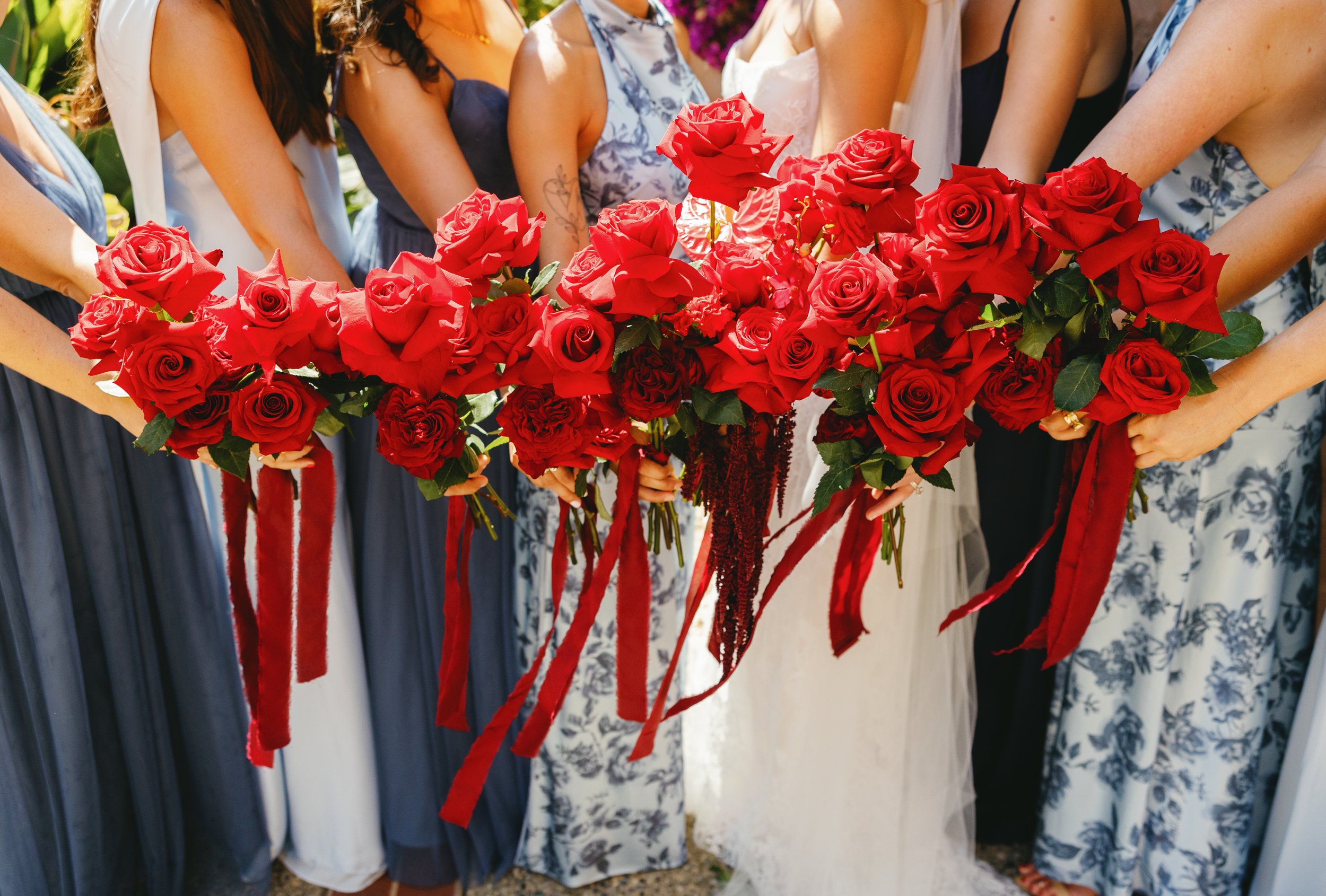 Bridesmaids holding lush red rose bouquets with flowing red ribbons during a Carmel Mission wedding ceremony.
