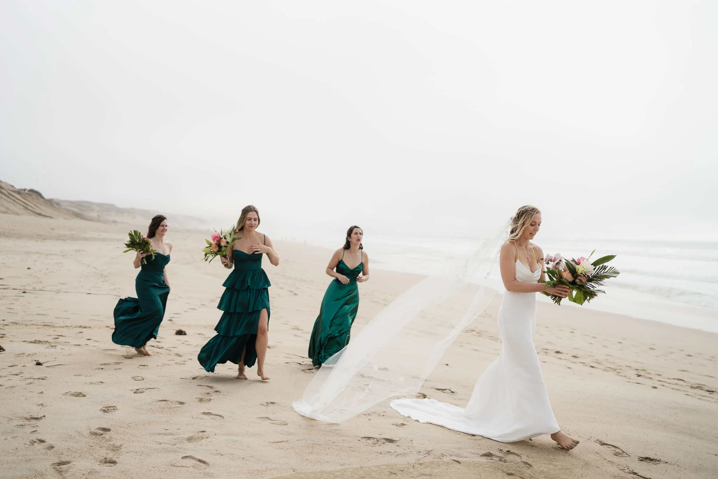 Bride in a white gown and flowing veil walks along a sandy beach carrying a bouquet, followed by three bridesmaids in teal dresses.