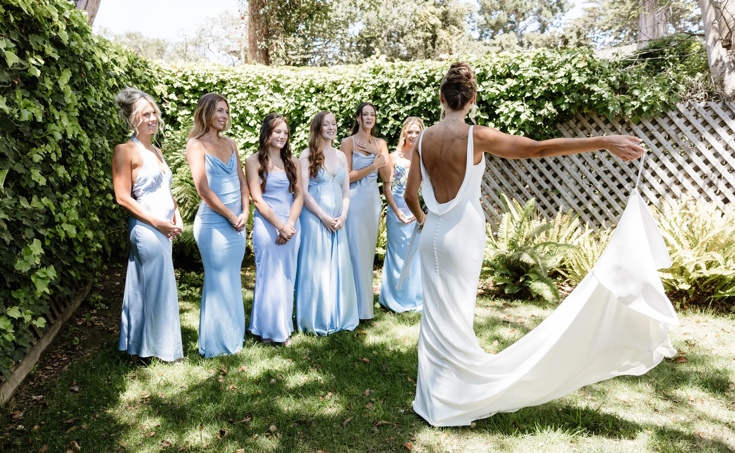 Bride in a low-back white wedding gown reveals her dress to bridesmaids wearing light blue dresses in a lush garden setting.