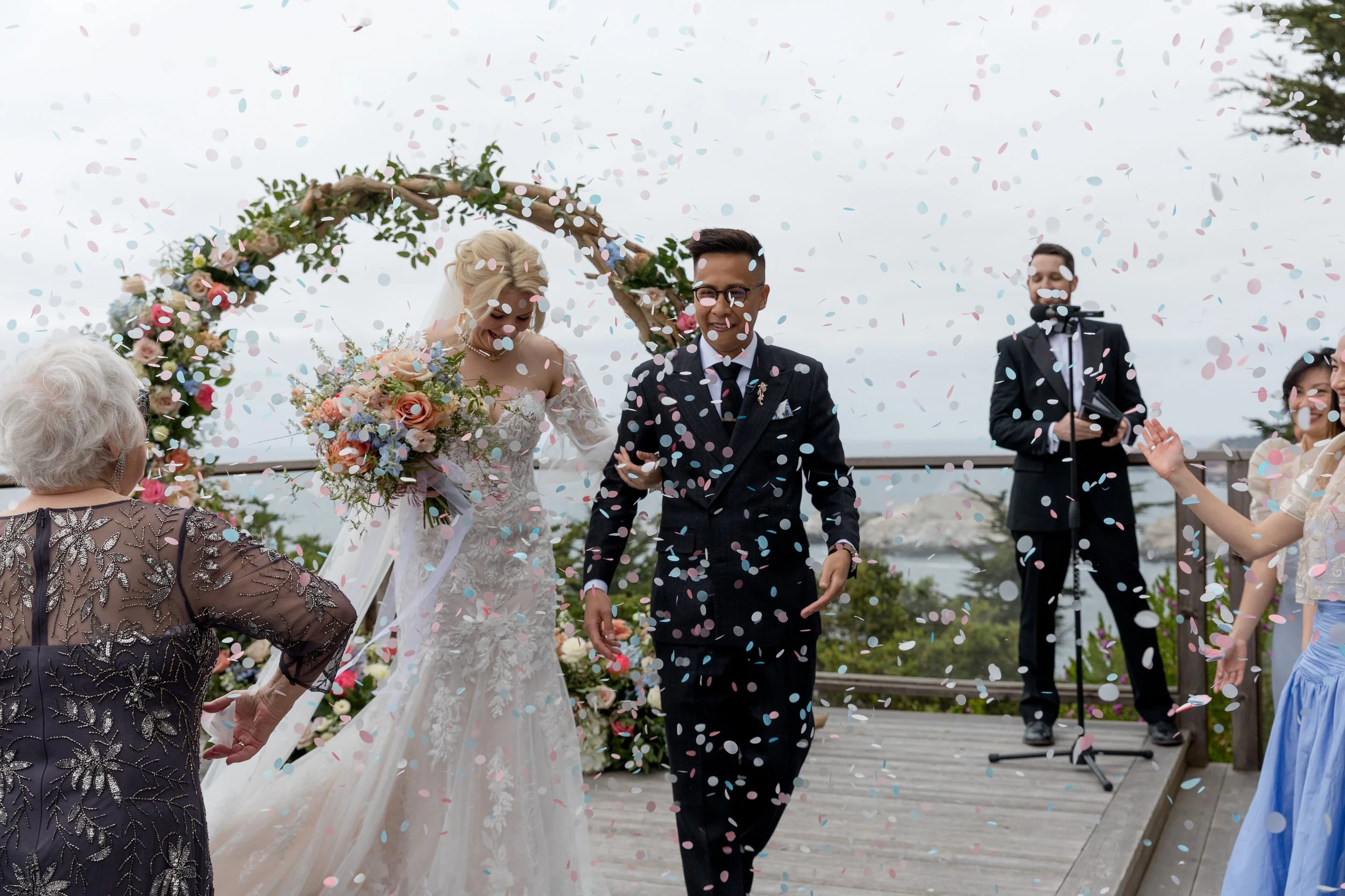Wedding couple walking through colorful confetti after an oceanfront ceremony with floral arch