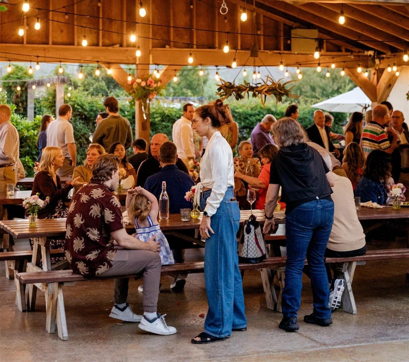 Outdoor gathering under string lights with people socializing at picnic tables.