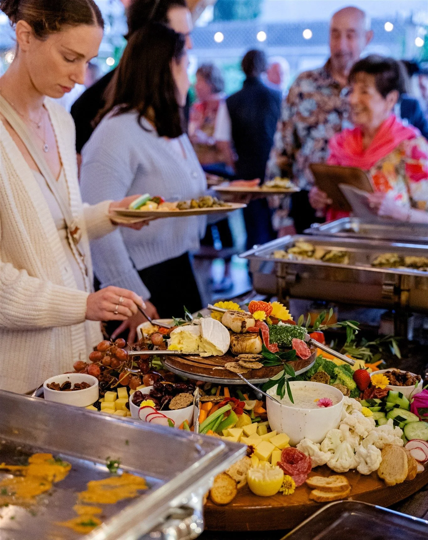 Guests at a buffet line serve themselves food from a table filled with cheese, fruit, vegetables, and appetizers at a social gathering.
