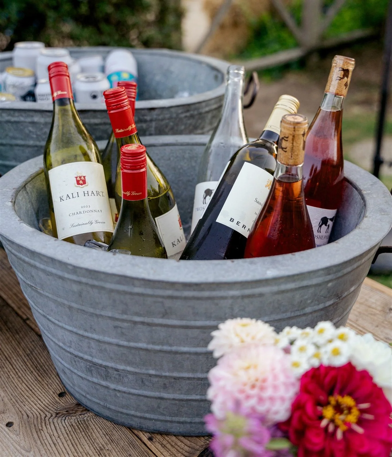 Several bottles of white, rosé, and red wine chilled in a large metal tub filled with ice, set on a wooden table with flowers in the foreground.