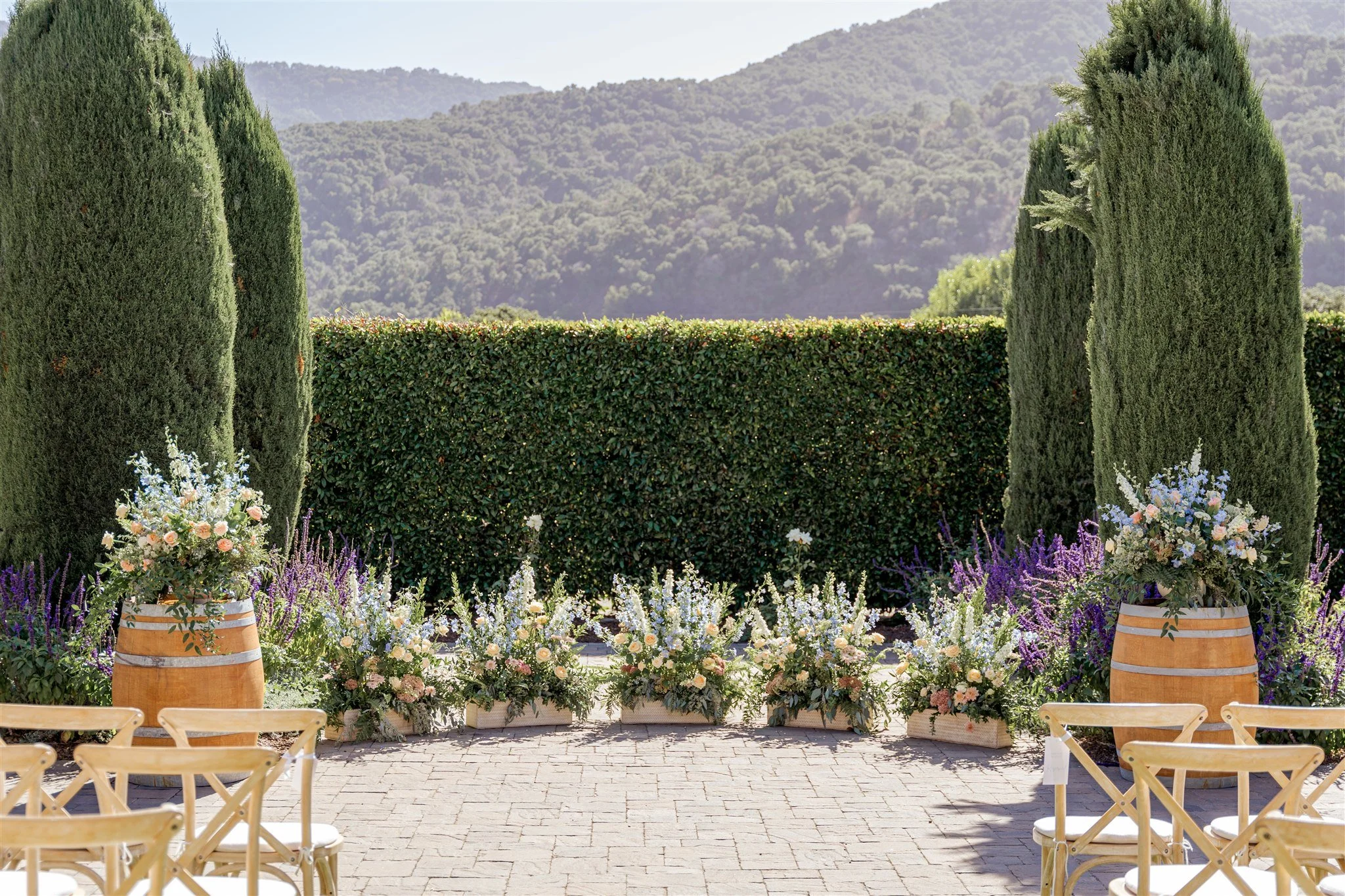 An outdoor wedding ceremony setup with wooden chairs facing a floral-lined aisle, featuring pastel flower arrangements in wooden planters and barrels, tall cypress trees, a manicured hedge, and forested hills in the background.